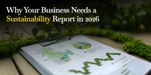 Blog post header featuring the title "Why Your Business Needs a Sustainability Report in 2026" in white and yellow text. The background displays a clipboard with green data visualization charts and graphs, resting on a wooden surface framed by fresh moss and greenery.