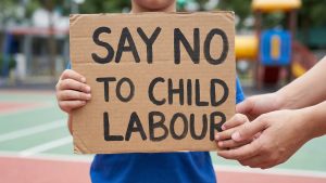 A close-up shot of a child's small hands holding a cardboard sign that reads "SAY NO TO CHILD LABOUR" in bold, hand-written black letters. The child is standing outdoors at a park or playground, and an adult’s hand is visible resting gently on the side of the sign in a gesture of support.